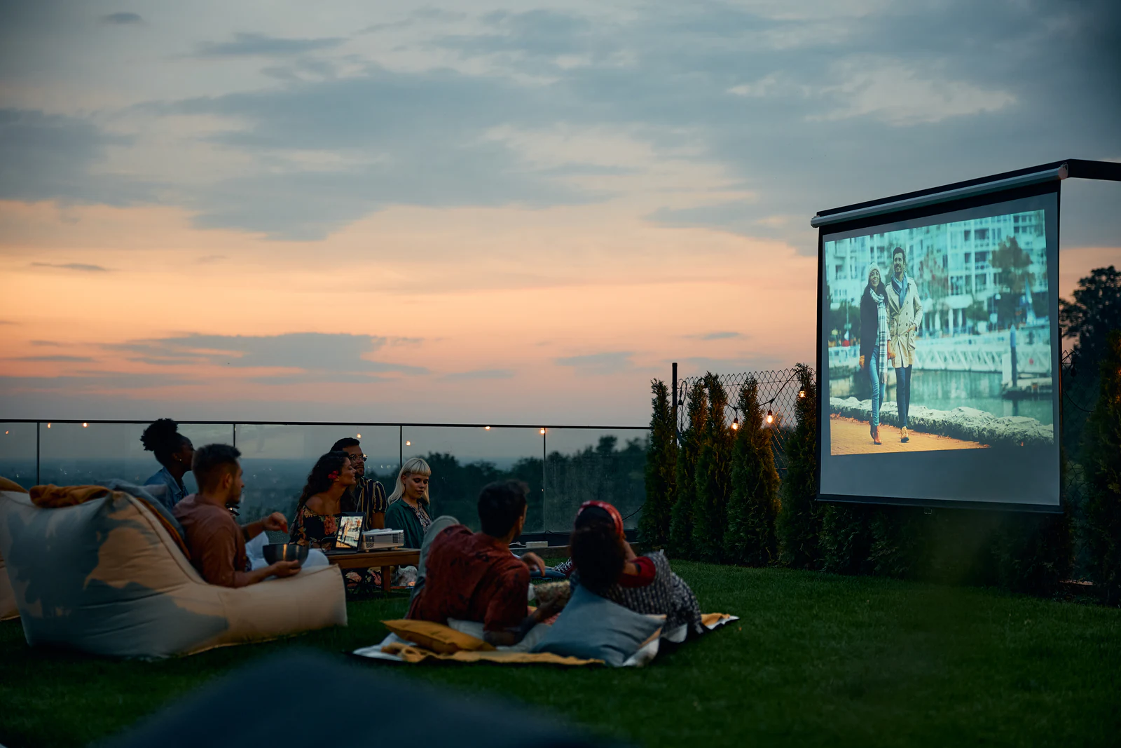 Grupa osób ogląda film na dużym ekranie podczas seansu plenerowego o zmierzchu